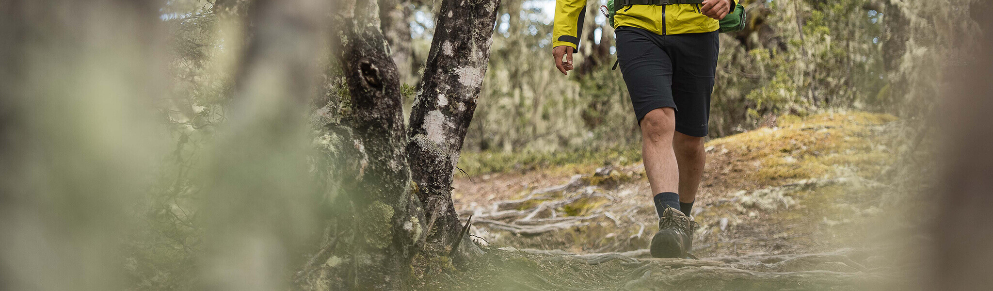 Man walking through forest wearing bright yellow jacket dark shorts and boots