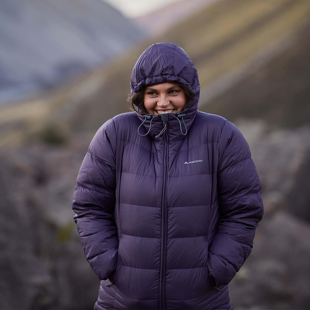 A woman wearing a dark purple macpac puffer jacket with the hood up, standing in a valley