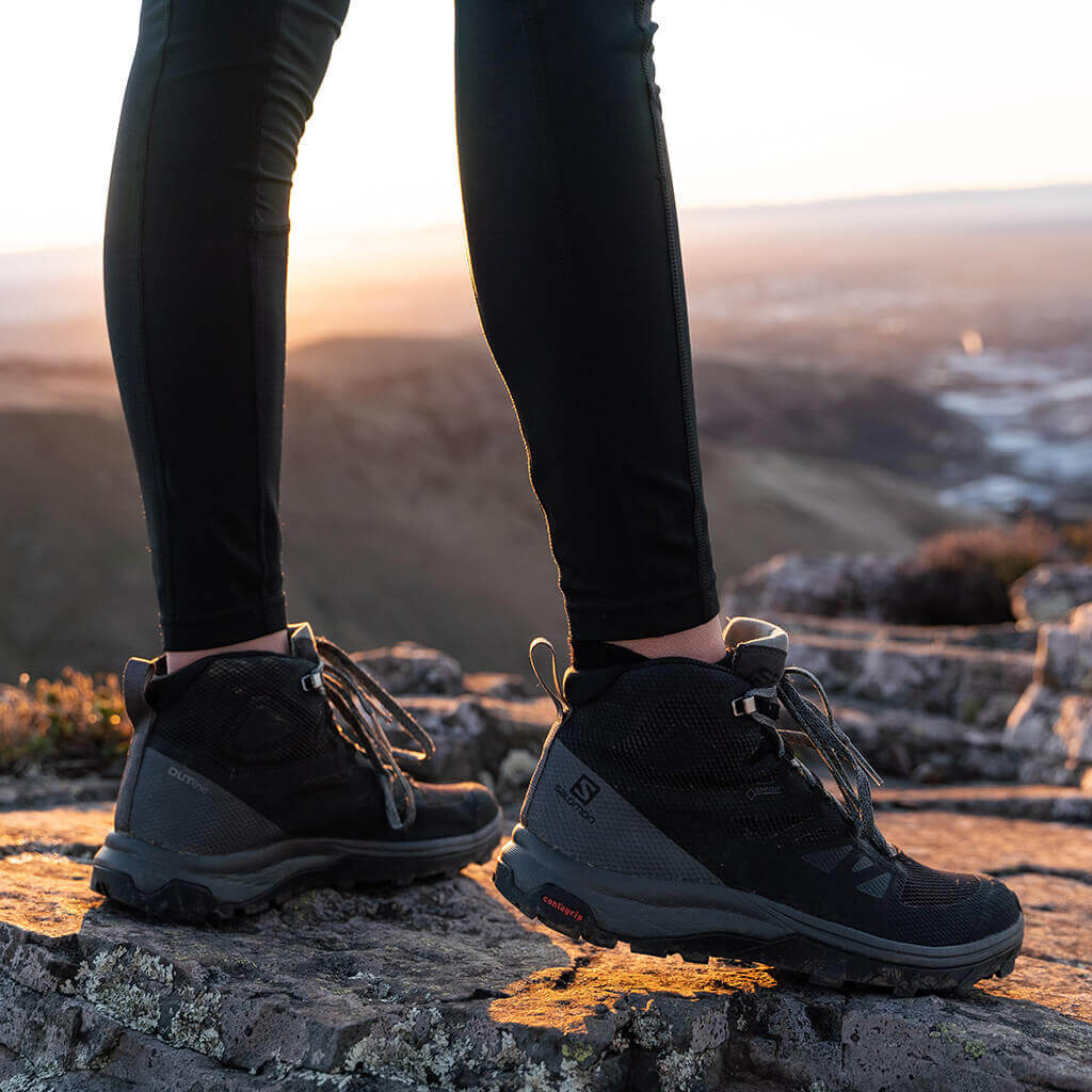 two people standing on side of river wearing hiking gear including boots
