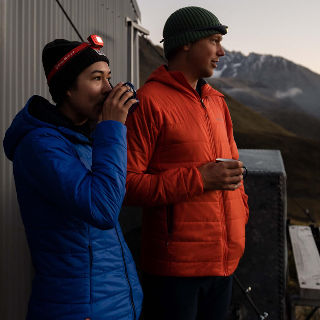 A man and a woman wearing hooded rain jackets hiking through a river using poles