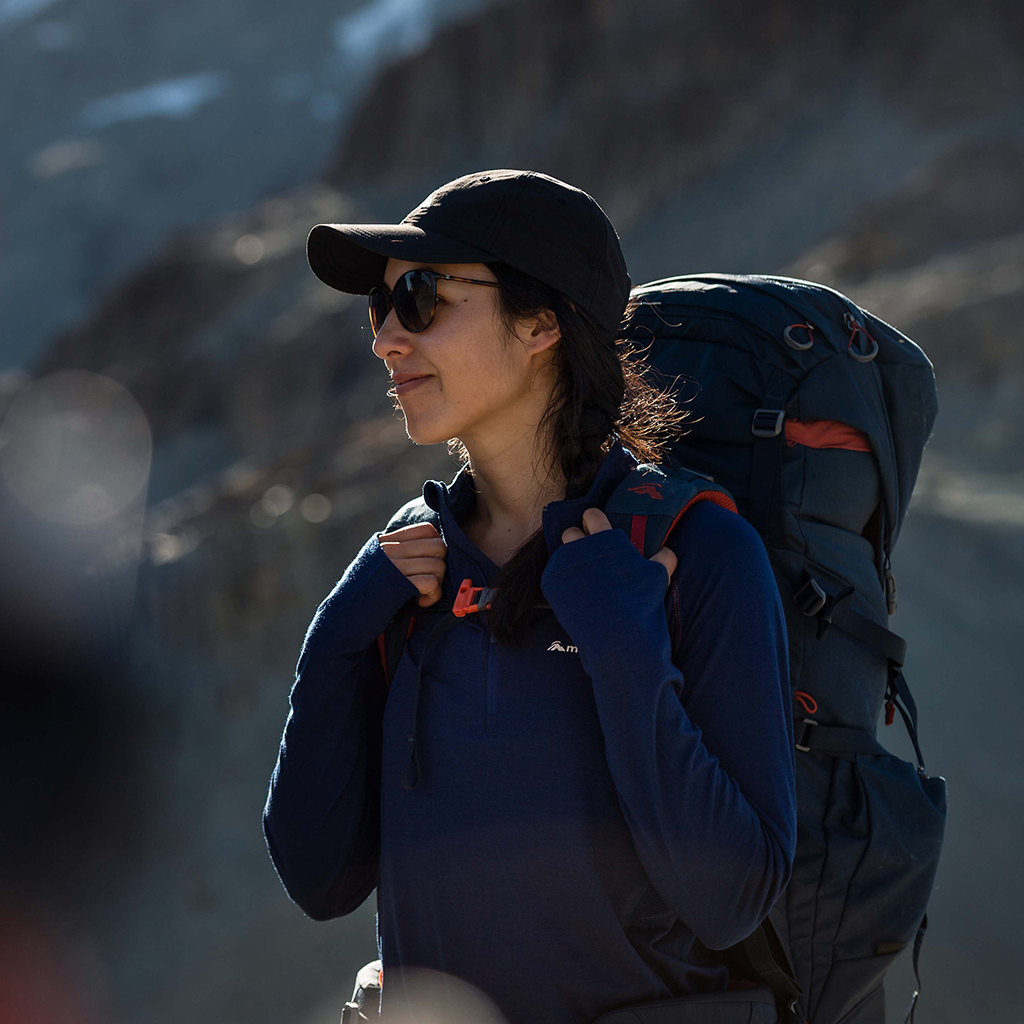 Woman wearing blue merino pullover standing next to a rocky cliff, wearing a backpack