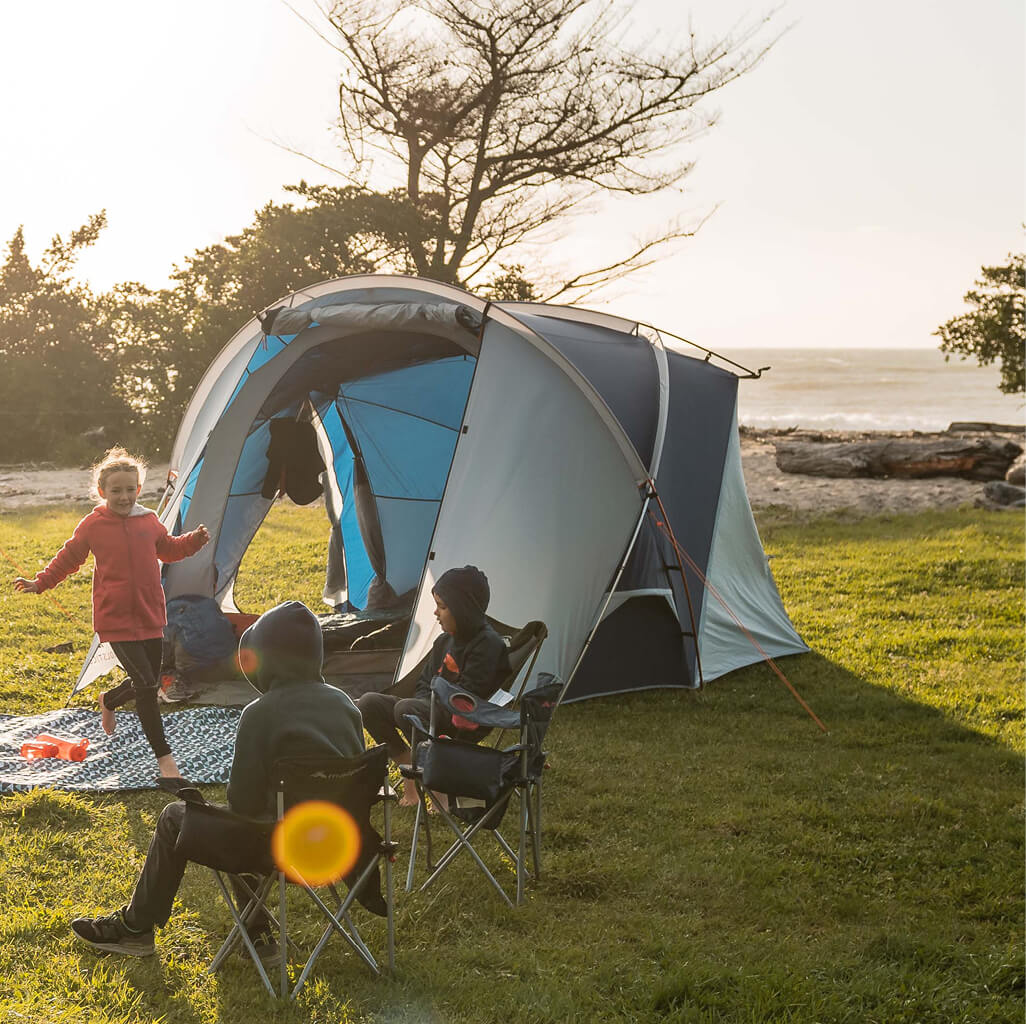 Three kids on grass outsite blue and grey Macpac tent pitched near a beach