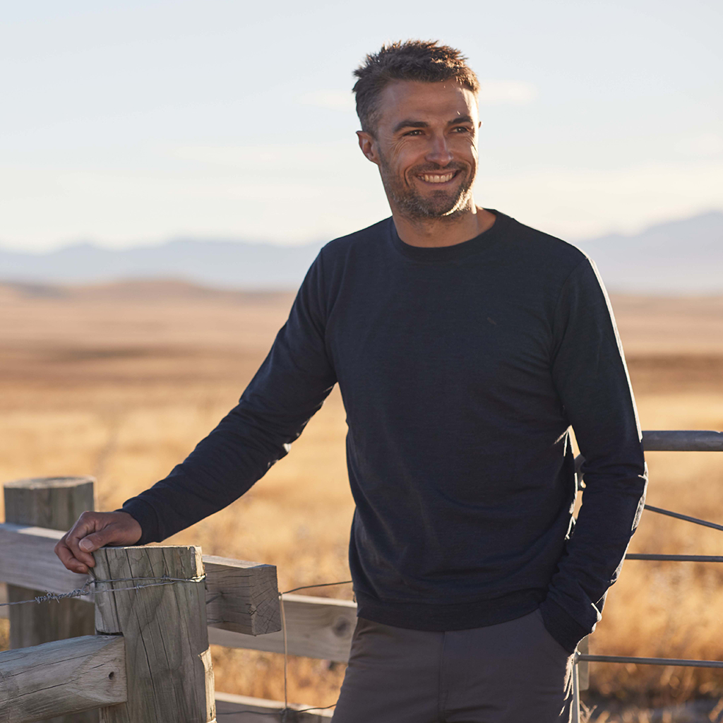 Man wearng dark merino top leaning on farm fence